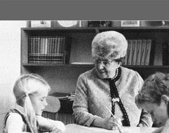 A black-and-white photo of Mae Carden in an elementary school classroom. She is writing on a piece of paper with a Number 2 pencil and tutoring a young girl.